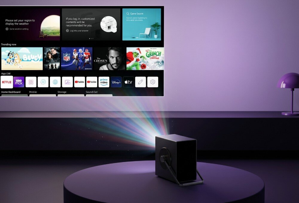 Na obrazie widać projektor wyświetlający interfejs z aplikacjami takimi jak Netflix, YouTube i Disney+. Projektor emituje kolorowe światło na ekran, a w tle znajduje się fioletowa lampa na stoliku.
