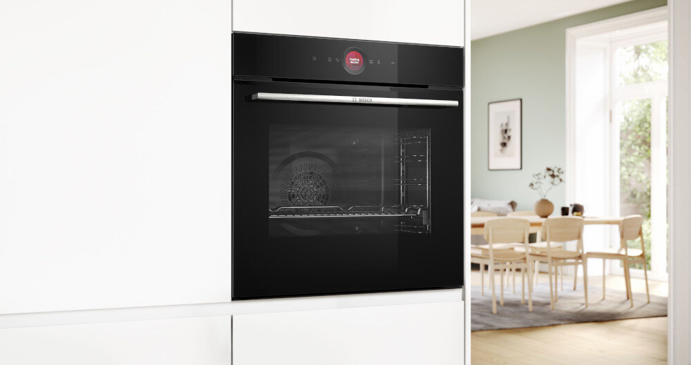 PIEKARNIK BOSCH HBG7242BB1 Czarny piekarnik jest wbudowany w białą zabudowę kuchenną, a w tle widać jasną jadalnię z drewnianym stołem i krzesłami. Front urządzenia odbija światło, podkreślając jego gładką powierzchnię do zabudowy zaawansowane funkcje intuicyjna obsługa programy pieczenia wyśmienite efekty komfort gotowania