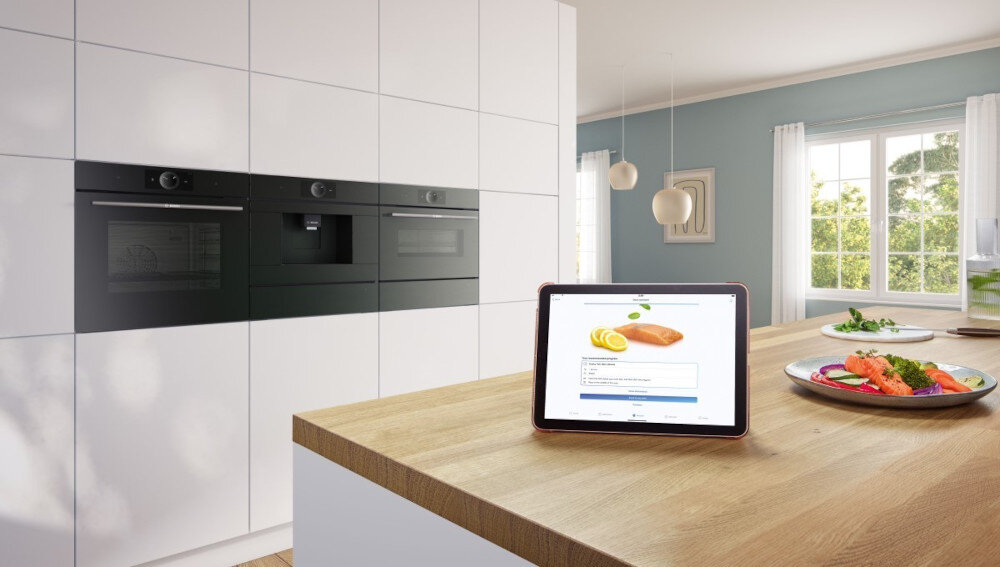 PIEKARNIK BOSCH HBG7242BB1 Na kuchennej wyspie stoi tablet z przepisem, a w tle znajduje się rząd trzech urządzeń do zabudowy w białej szafkowej ścianie. Obok leży talerz z kolorowym daniem, tworząc scenę gotowania z wykorzystaniem technologii 
