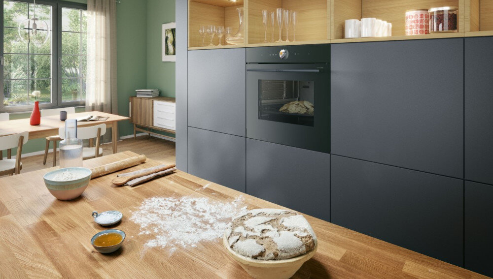 PIEKARNIK BOSCH HBG7242BB1 Na blacie kuchennym leży miska z wyrobionym ciastem, obok rozsypana jest mąka, wałek oraz składniki potrzebne do pieczenia. W zabudowie po drugiej stronie pomieszczenia piekarnik podgrzewa chleb, tworząc ciepłą, domową atmosferę Home Connect aplikacja nowinki kucharskie przepisy porady