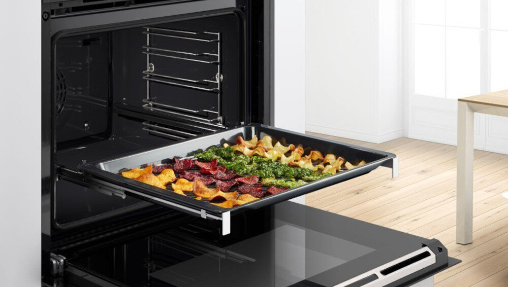 PIEKARNIK BOSCH HBG7242BB1 Z piekarnika wysunięta jest blacha z warzywami w kilku kolorach, ułożonymi w oddzielnych sekcjach. W tle widać jasną kuchnię z dużym oknem, co nadaje scenie domowy charakter Air Fry zdrowe pieczenie mniej tłuszczu