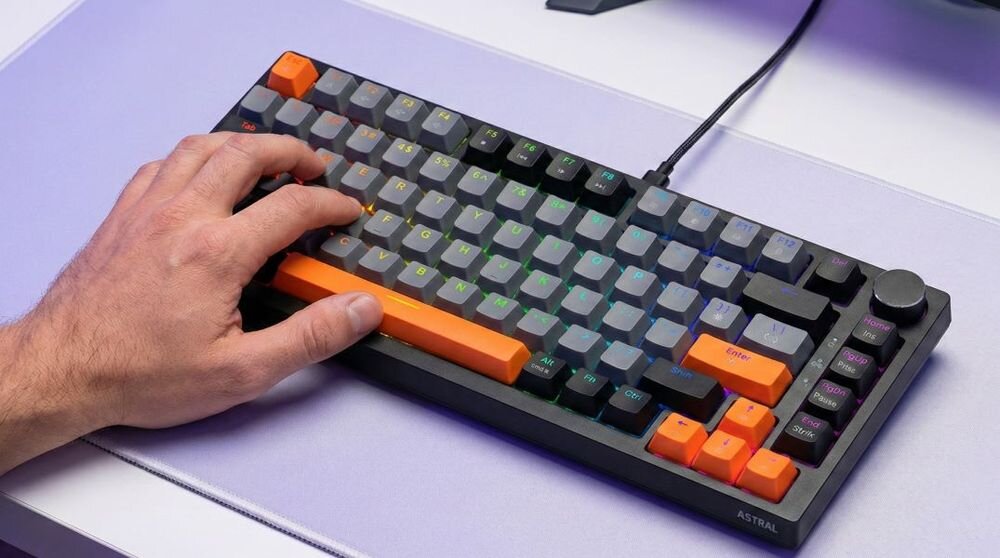 Klawiatura mechaniczna z czarno-szaro-pomarańczowymi klawiszami, z podświetleniem RGB. Na klawiaturze widoczna jest dłoń, a na obudowie napis \'ASTRAL\'.