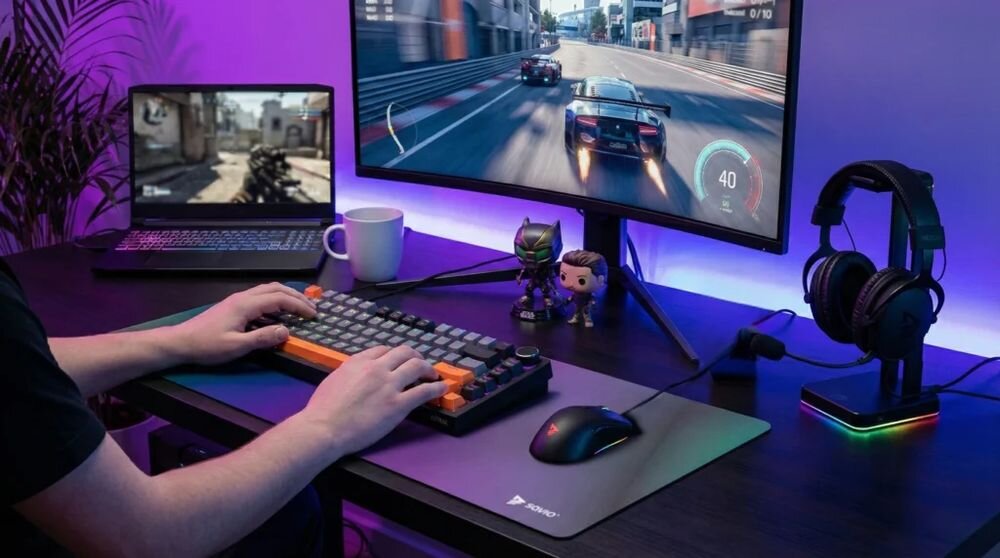 Osoba używa klawiatury o czarno-szaro-pomarańczowym wzorze przy biurku z dwoma monitorami, na których wyświetlane są gry. Na biurku znajdują się także słuchawki na stojaku, kubek oraz dwie figurki.