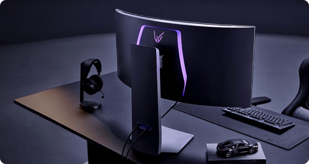Monitor LG UltraGear 34GS95QE-B.AEU Widok monitora od tyłu na eleganckim, czarnym biurku. Z tyłu obudowy widać subtelne podświetlenie LED w kształcie sześciokąta, podstawa, regulacja, wysokość, nachylenie, montaż VESA