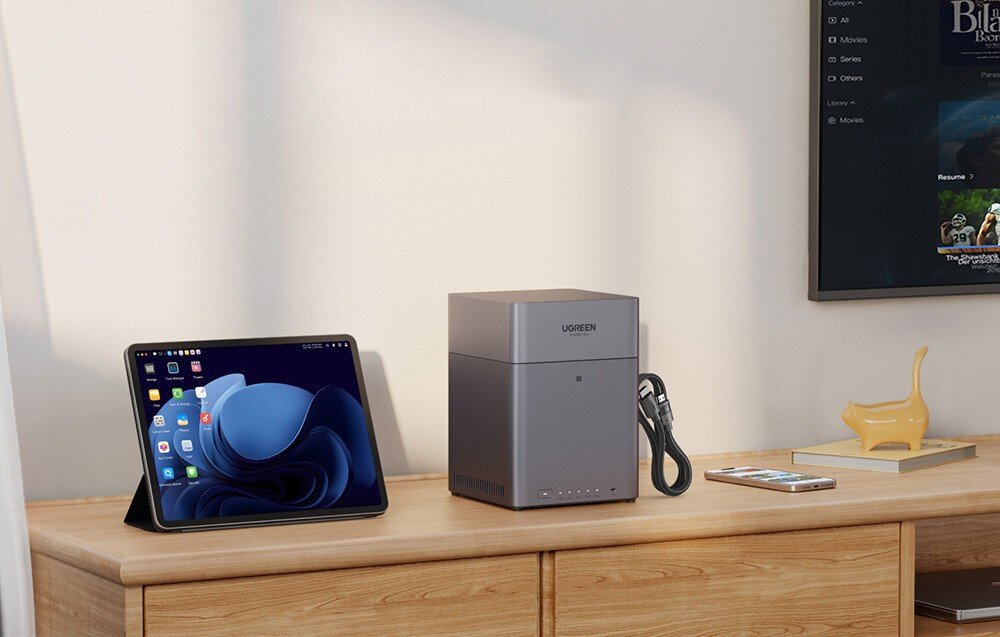 Na drewnianej powierzchni stoi szary serwer plików z logo \'UGREEN\', obok którego znajduje się tablet z wyświetlonym ekranem głównym. W tle widać fragment telewizora z interfejsem użytkownika.