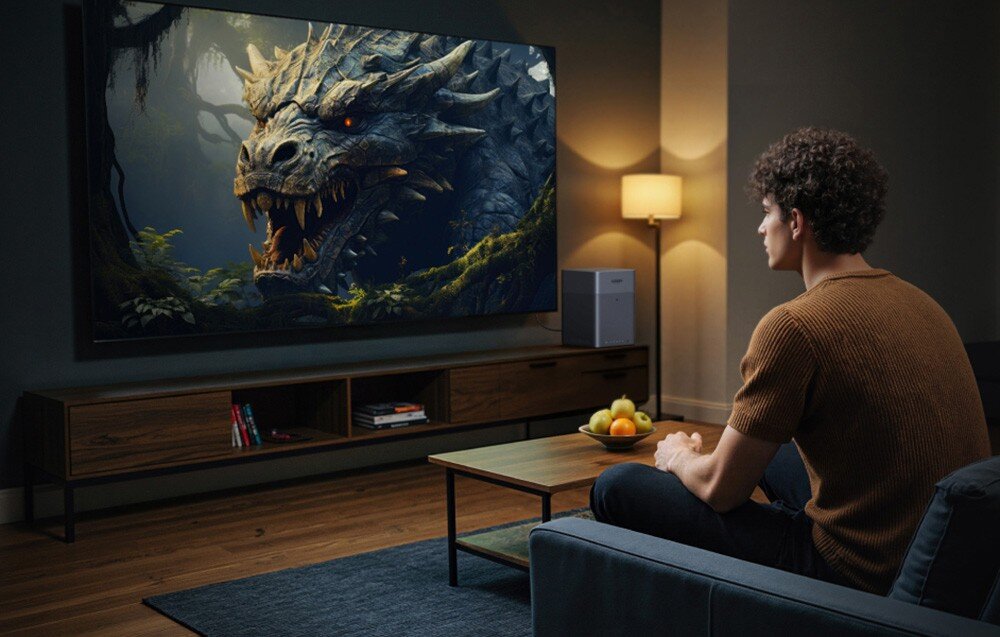 Osoba siedzi na kanapie w salonie, oglądając na dużym telewizorze obraz przedstawiający smoka w lesie. Obok telewizora znajduje się lampa stojąca i niewielkie urządzenie elektroniczne.
