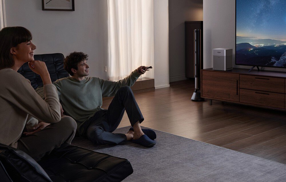 Dwie osoby siedzą w salonie, patrząc na duży telewizor wyświetlający krajobraz górski. Obok telewizora znajduje się drewniana szafka z urządzeniem elektronicznym na blacie.