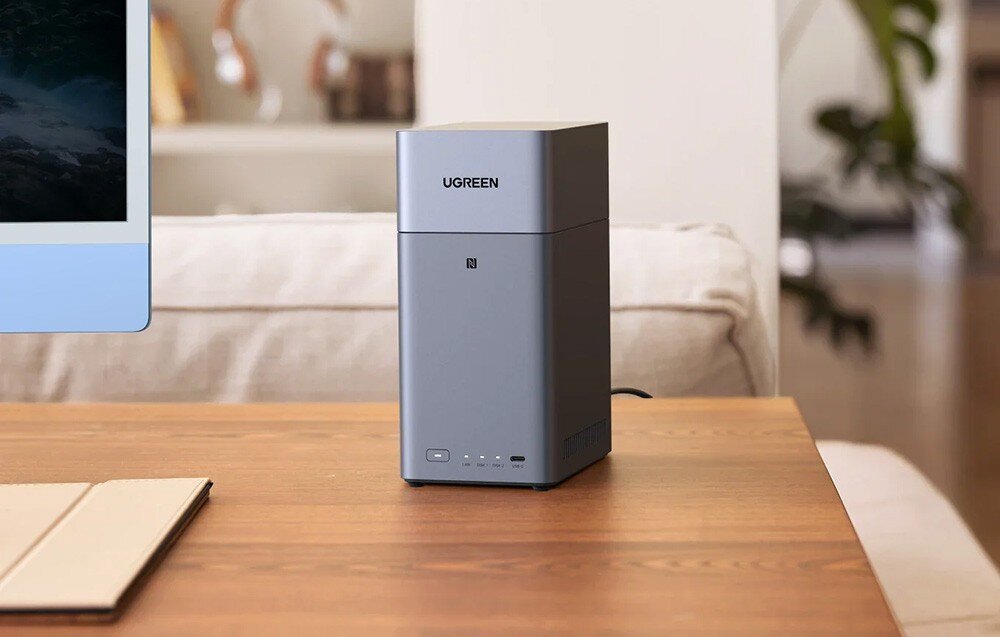 Na drewnianym stole stoi szary serwer plików z napisem \'UGREEN\' na górze. W tle widoczny jest fragment monitora i jasna sofa.