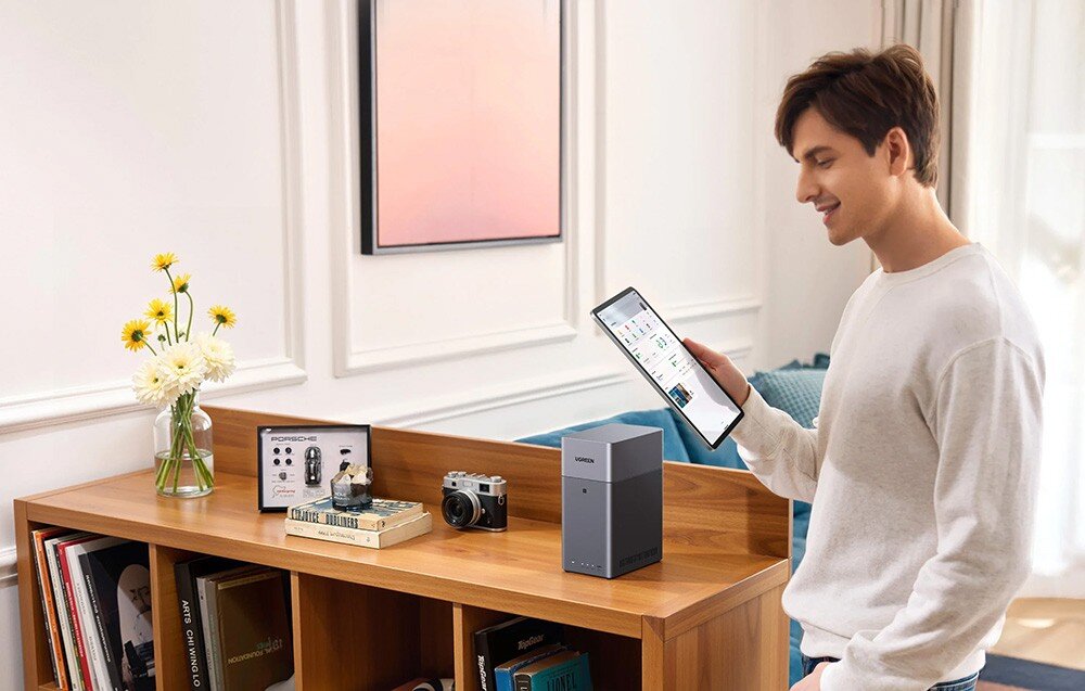 Na drewnianej półce stoi serwer plików z logo UGREEN. Obok znajduje się aparat fotograficzny i pudełko z napisem \'Porsche Design\'.