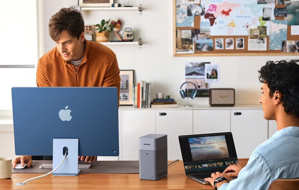 Dwóch mężczyzn pracuje przy biurku, na którym znajduje się komputer stacjonarny z dużym niebieskim monitorem oraz laptop. Na biurku stoi szary serwer plików z napisem \'UGREEN\'.