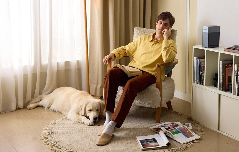 Mężczyzna siedzi na fotelu z książką, obok leży pies na dywanie. Na półce znajduje się urządzenie przypominające serwer plików oraz kilka książek.