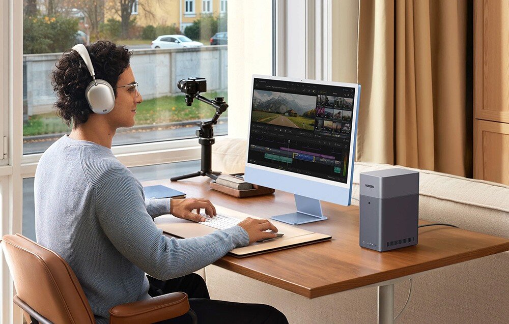 Osoba siedzi przy biurku z komputerem, na którym wyświetlany jest program do edycji wideo. Obok monitora znajduje się szary serwer plików z napisem \'UGREEN\'.