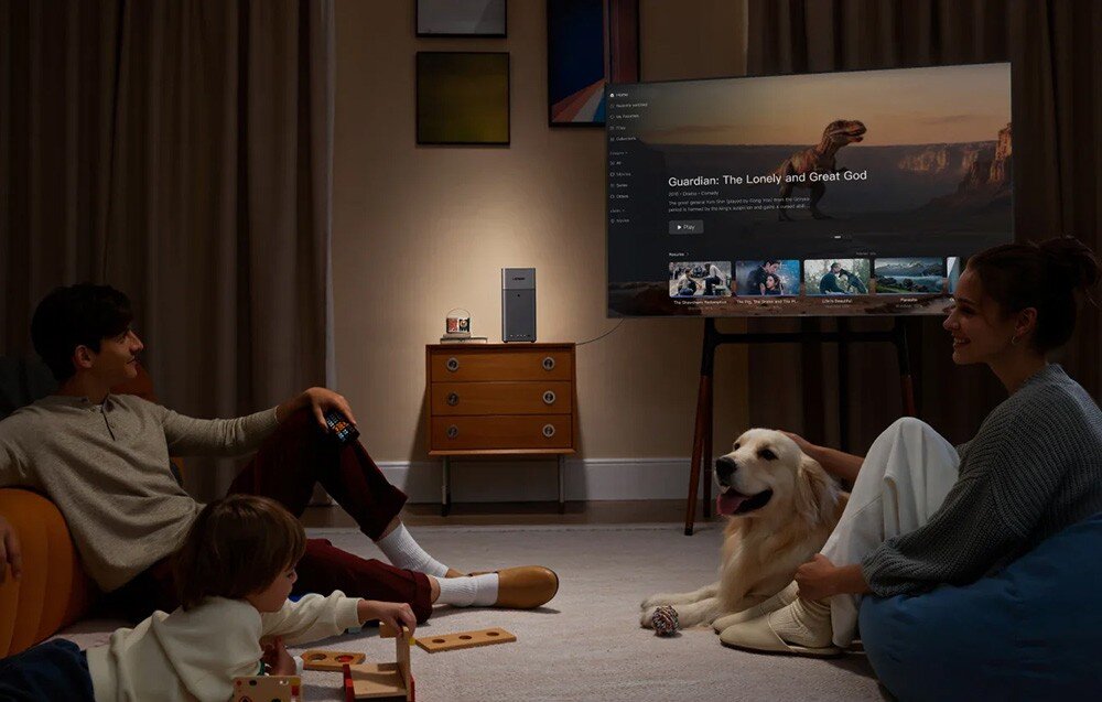 W salonie znajduje się telewizor wyświetlający interfejs z filmem \'Guardian: The Lonely and Great God\'. Na szafce obok telewizora stoi urządzenie przypominające serwer plików.