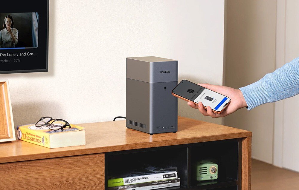 Na drewnianej półce stoi szary serwer plików z logo UGREEN. Obok osoba trzyma smartfon z wyświetlonym ekranem aplikacji.