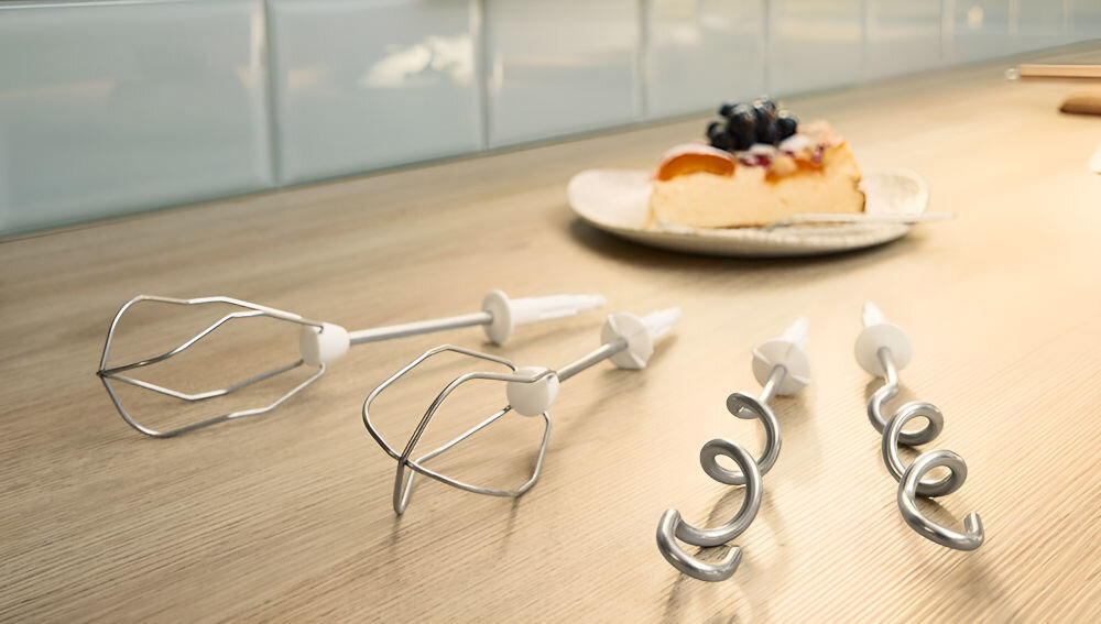 Na drewnianym blacie leżą cztery metalowe końcówki do miksera: dwie trzepaczki i dwa haki do ciasta. W tle na talerzu znajduje się kawałek ciasta z owocami.