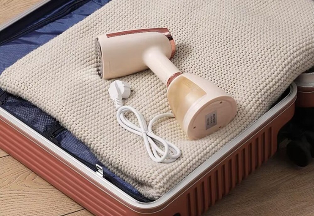 Na obrazie widoczna jest parownica do ubrań w kolorze beżowym, leżąca na złożonym swetrze w walizce. Obok parownicy znajduje się zwinięty biały kabel z wtyczką.