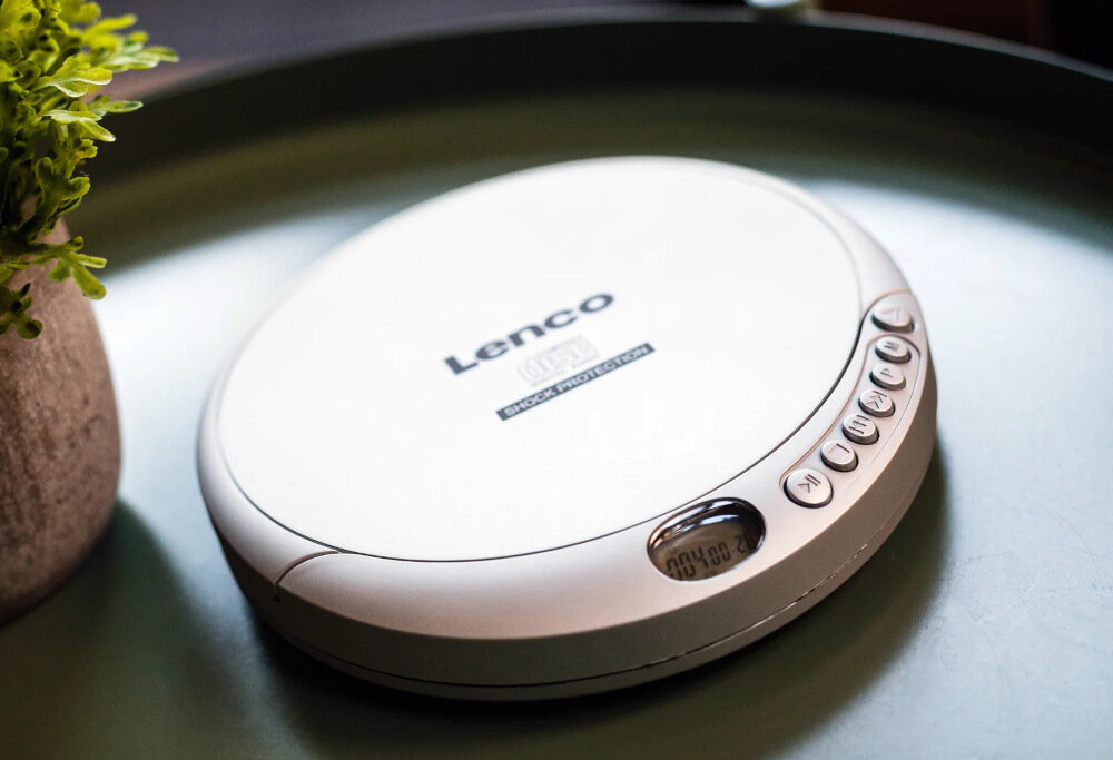 Srebrny odtwarzacz CD z logo \'Lenco\' na górze, posiada przyciski sterujące i mały wyświetlacz LCD. Obok znajduje się zielona roślina w doniczce.