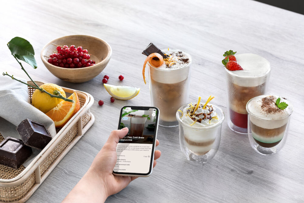 Smartfon z otwartą aplikacją De'Longhi prezentującą przepisy na kawy mrożone z owocami i czekoladą obok gotowych napojów.