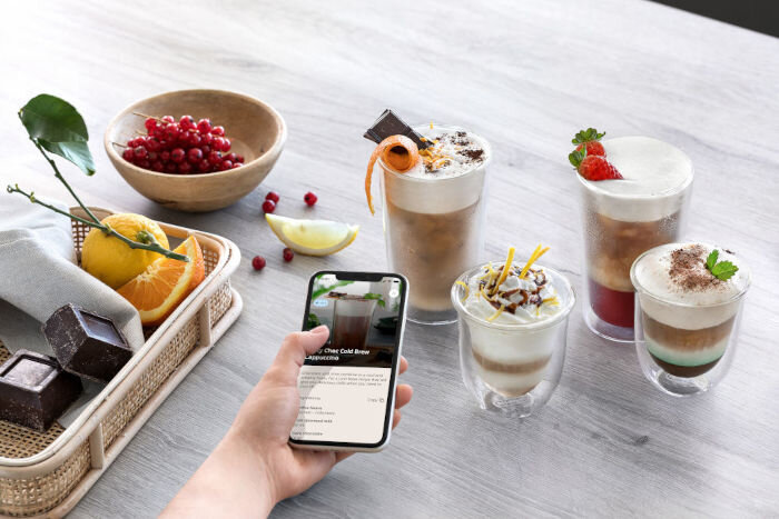 Smartfon z otwartą aplikacją De'Longhi prezentującą przepisy na kawy mrożone z owocami i czekoladą obok gotowych napojów.