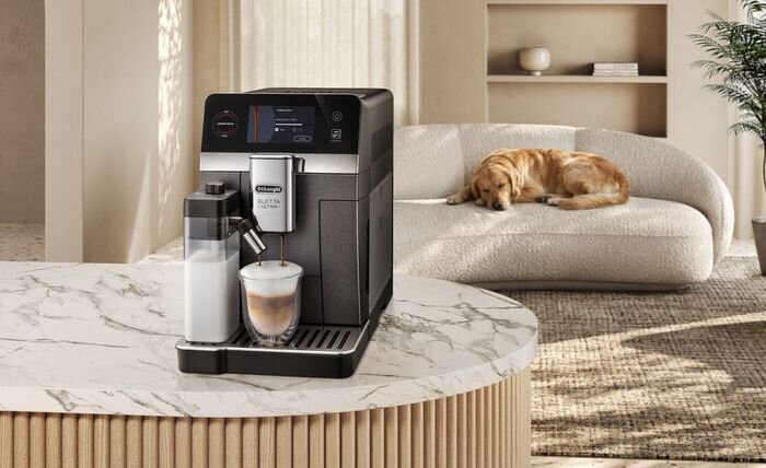 Na marmurowym blacie stoi ekspres do kawy z logo \'DeLonghi\', obok którego znajduje się szklanka z kawą i pojemnik na mleko. W tle widać kanapę, na której leży pies.