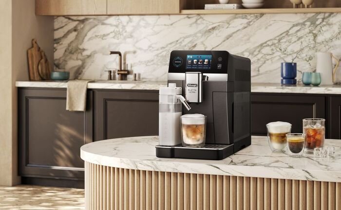 Na marmurowym blacie kuchennym stoi ekspres do kawy z logo \'DeLonghi Eletta\'. Obok ekspresu znajdują się szklanki z różnymi napojami kawowymi.