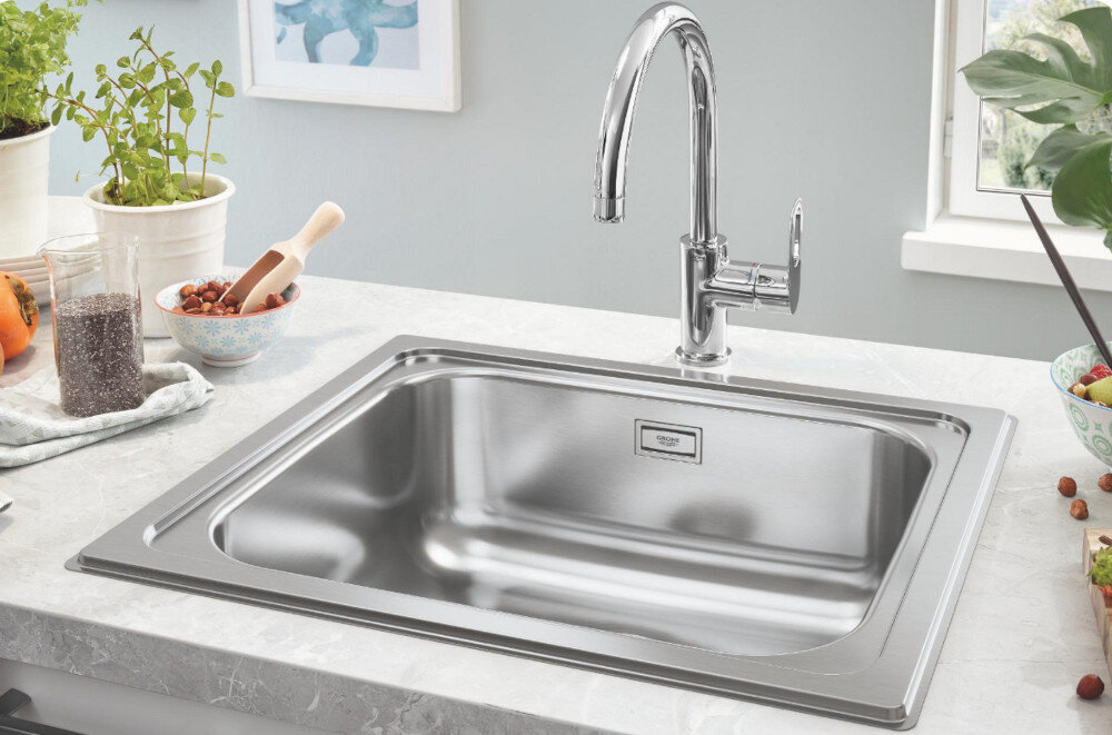 Zlewozmywak jednokomorowy GROHE K200 31719SD0 Stal szlachetna Na zdjęciu widać stalowy zlewozmywak jednokomorowy z ociekaczem, osadzony w blacie o ciemnoszarej powierzchni. W centralnej części ociekacza znajduje się chromowana, jednouchwytowa bateria stojąca, a w tle widoczne są minimalistyczne akcesoria kuchenne na białej półce.