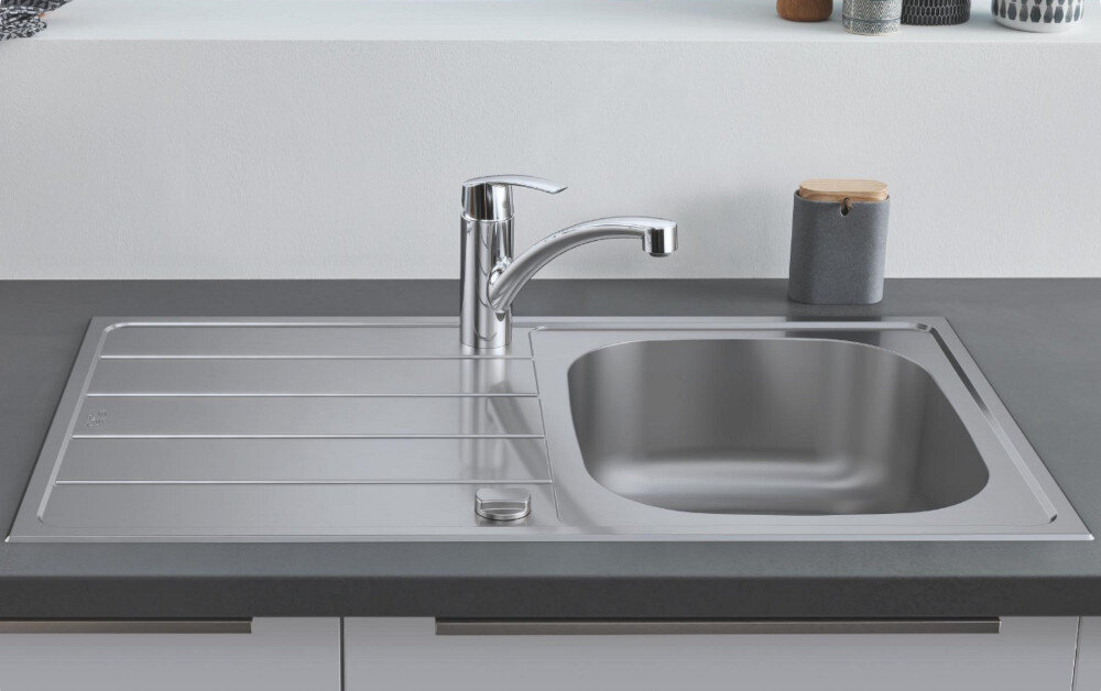 Zlewozmywak jednokomorowy z ociekaczem GROHE K200 31552SD1 Stal szlachetna Na zdjęciu widać stalowy zlewozmywak jednokomorowy z ociekaczem, osadzony w blacie o ciemnoszarej powierzchni. W centralnej części ociekacza znajduje się chromowana, jednouchwytowa bateria stojąca, a w tle widoczne są minimalistyczne akcesoria kuchenne na białej półce.