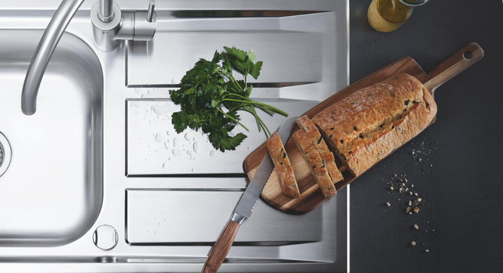 Zlewozmywak jednokomorowy z ociekaczem GROHE K200 31552SD1 Stal szlachetna Na zdjęciu z góry widać stalowy zlewozmywak z ociekaczem, na którym leżą świeże gałązki pietruszki i krople wody. Obok, na drewnianej desce do krojenia ułożonej na ociekaczu, znajduje się pokrojony bochenek chleba z nożem o drewnianej rękojeści, a na blacie widać rozsypane okruchy i butelkę z oliwą stal nierdzewna materiał wykonania wyjątkowa wytrzymałość odporny na korozję uszkodzenia mechaniczne odkształcania