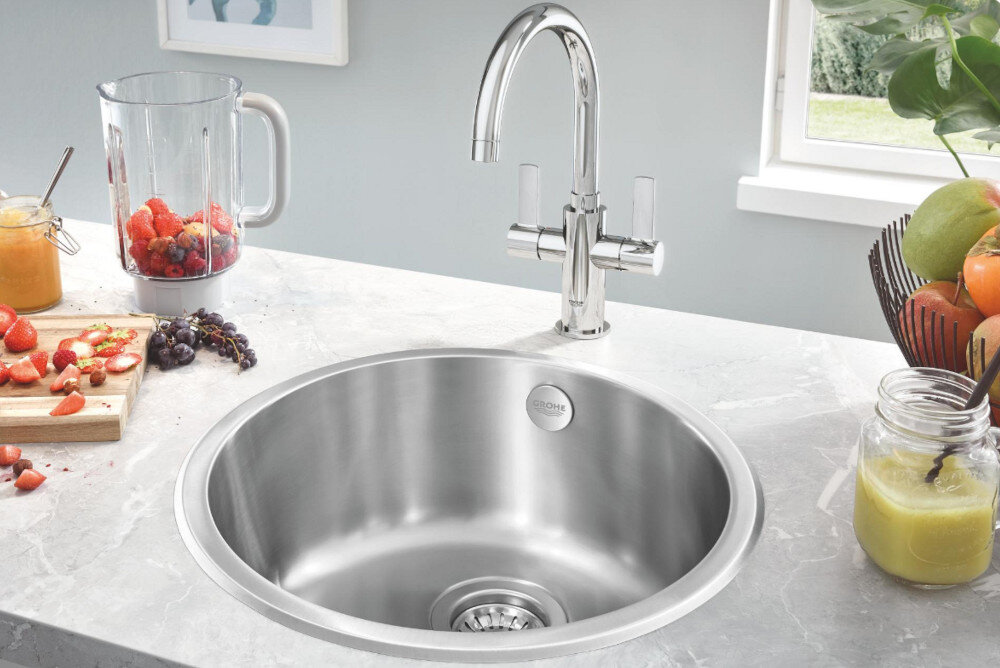 Zlewozmywak jednokomorowy GROHE K200 31720SD0 Stal szlachetna Na zdjęciu widać stalowy zlewozmywak jednokomorowy z ociekaczem, osadzony w blacie o ciemnoszarej powierzchni. W centralnej części ociekacza znajduje się chromowana, jednouchwytowa bateria stojąca, a w tle widoczne są minimalistyczne akcesoria kuchenne na białej półce.