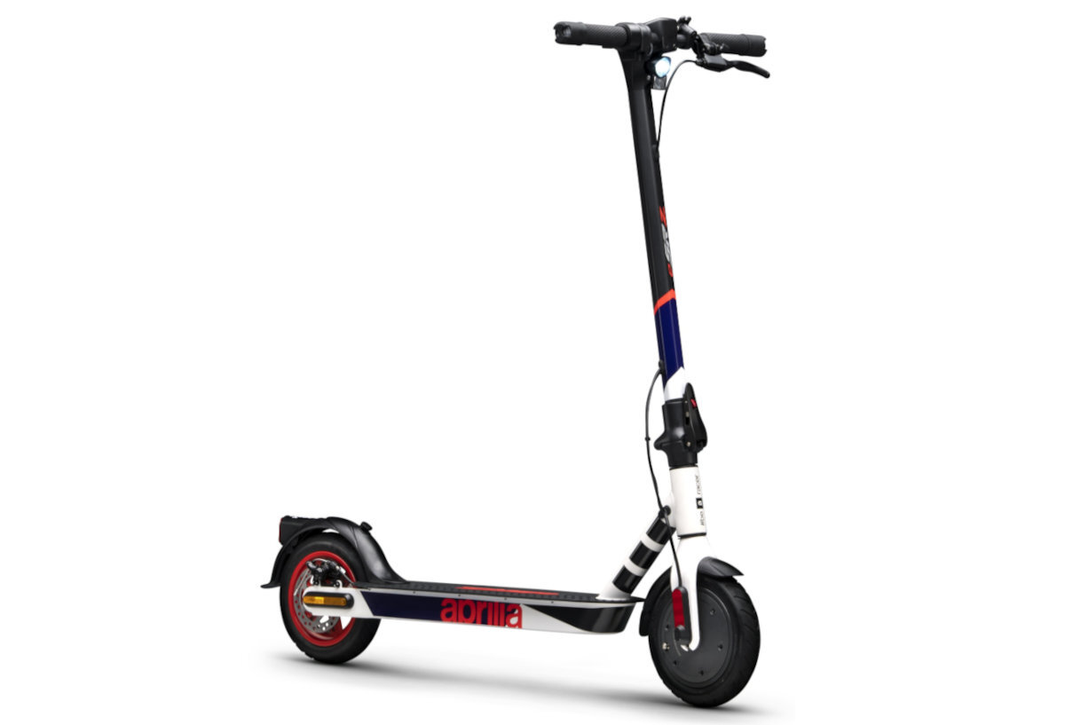 Hulajnoga elektryczna w kolorach biało-czarno-czerwonym z widocznym napisem \'aprilia\' na boku. Posiada duże koła i prostą kierownicę z hamulcem ręcznym.