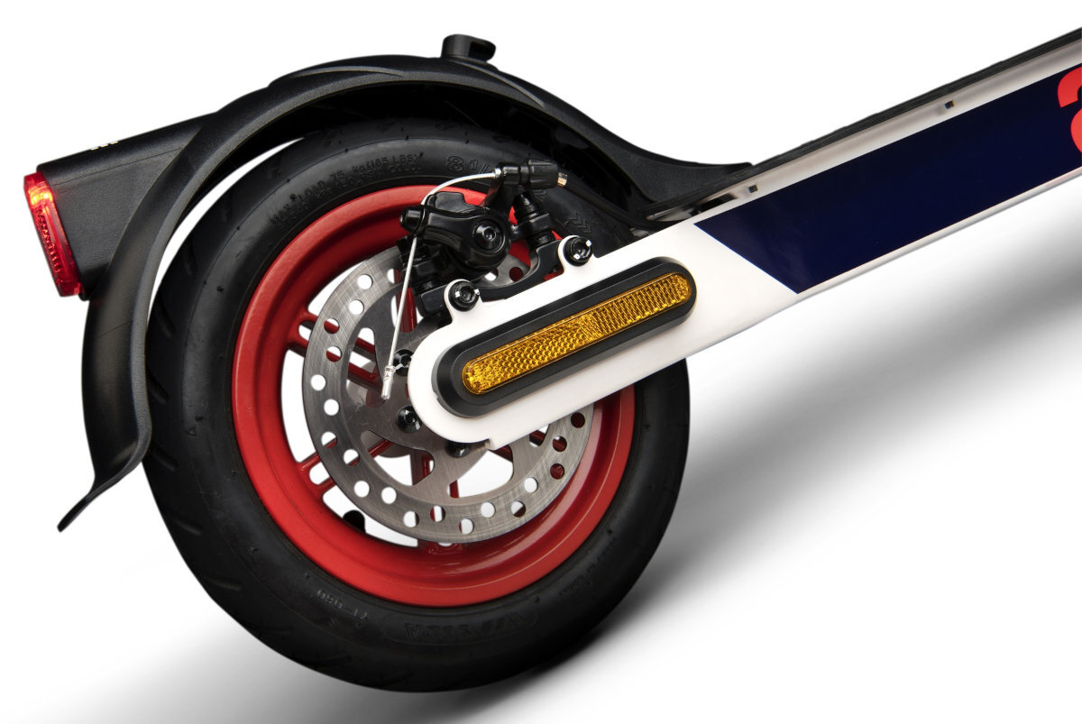 Zdjęcie przedstawia tylne koło hulajnogi elektrycznej z czerwonym obręczą i tarczą hamulcową. Na boku widoczny jest biały element z żółtym odblaskiem.
