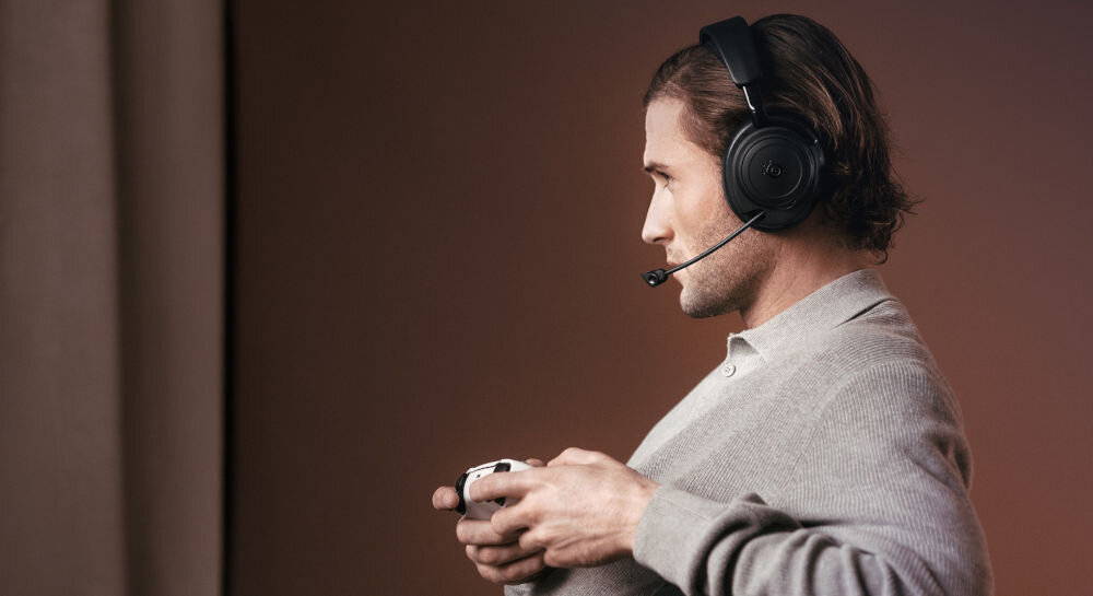 Słuchawki STEELSERIES Arctis Nova Elite metalowa konstrukcja komfort aplikacja mobilna presety gier, osoba z założonymi słuchawkami nausznymi z mikrofonem trzymająca kontroler w ręku
