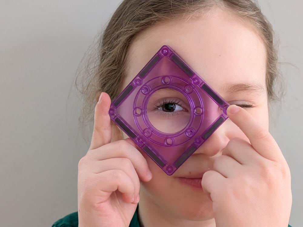 Klocki magnetyczne ASKATO Genialny dzieciak 127705 Dziewczynka patrząca przez okrągły otwór w fioletowym, kwadratowym klocku magnetycznym.