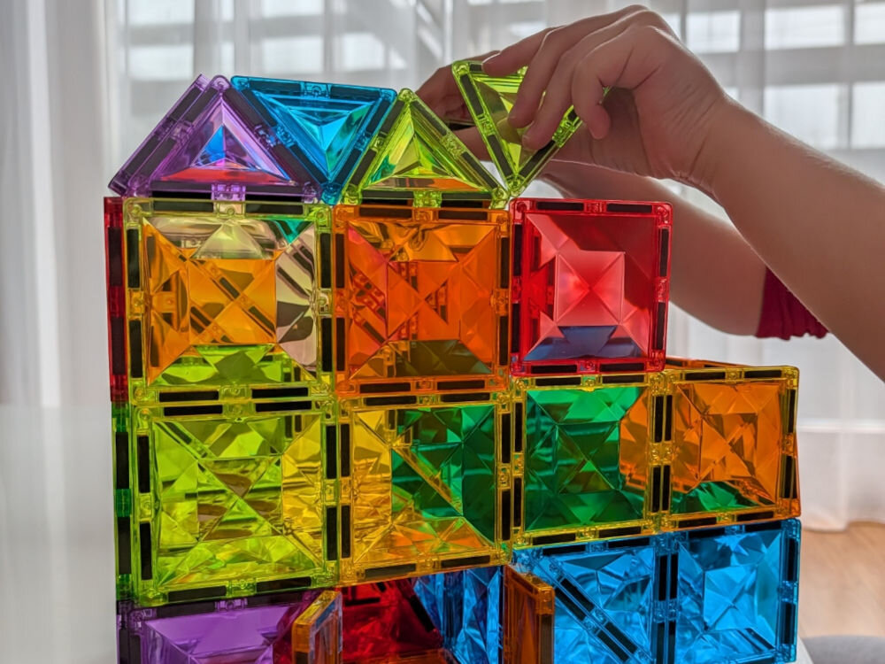 Klocki magnetyczne ASKATO Genialny dzieciak 127712 Wielokolorowy Ręce dziecka budującego konstrukcję 3D z przeźroczystych, kolorowych klocków magnetycznych przy oknie.