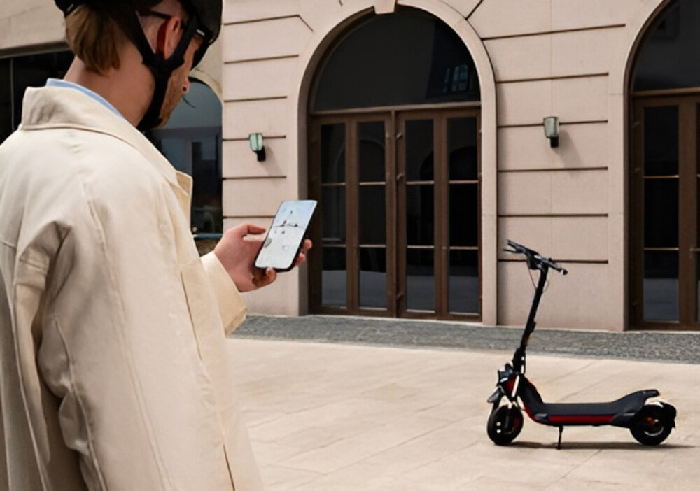 Hulajnoga elektryczna SEGWAY Ninebot ZT3 Pro D 70km 1600W Amortyzacja 11 cali Czarny miasto osoba stoi przed budynkiem trzyma telefon w dłoni przed budynkiem stoi hulajnoga wbudowany moduł Bluetooth aplikacja mobilna Segway-Ninebot Smart App na systemy Android i iOS