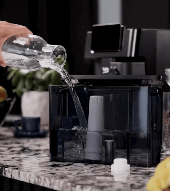 Osoba nalewa wodę z dzbanka do zbiornika ekspresu do kawy. W tle widoczny jest ekspres do kawy z logo Beko.