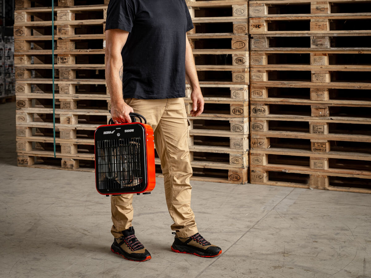 Nagrzewnica elektryczna DEDRA DED9922B, lekka, 6kg, uchwyt, ergonimia, Osoba niesie DEDRA DED9922B za uchwyt, nagrzewnica mobilna gotowa do transportu w hali.