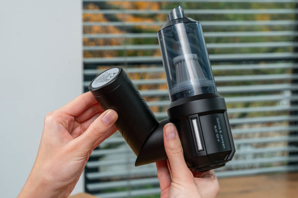 Odkurzacz ręczny GÖTZE & JENSEN HV400 Kompaktowy, czarny odkurzacz ręczny Götze & Jensen z przezroczystym pojemnikiem na kurz, trzymany przez dłonie. Widoczne jest gniazdo na filtr oraz logo marki.