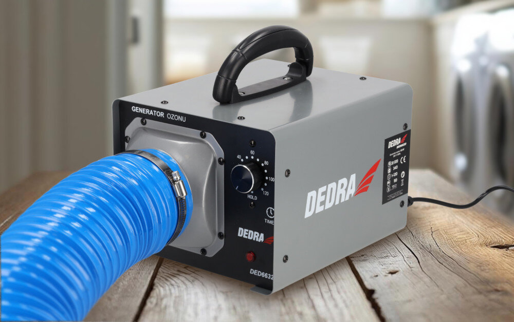 Na drewnianym stole znajduje się generator ozonu z napisem \'DEDRA\' i \'GENERATOR OZONU\'. Urządzenie ma szary kolor, czarny uchwyt i podłączony niebieski wąż.