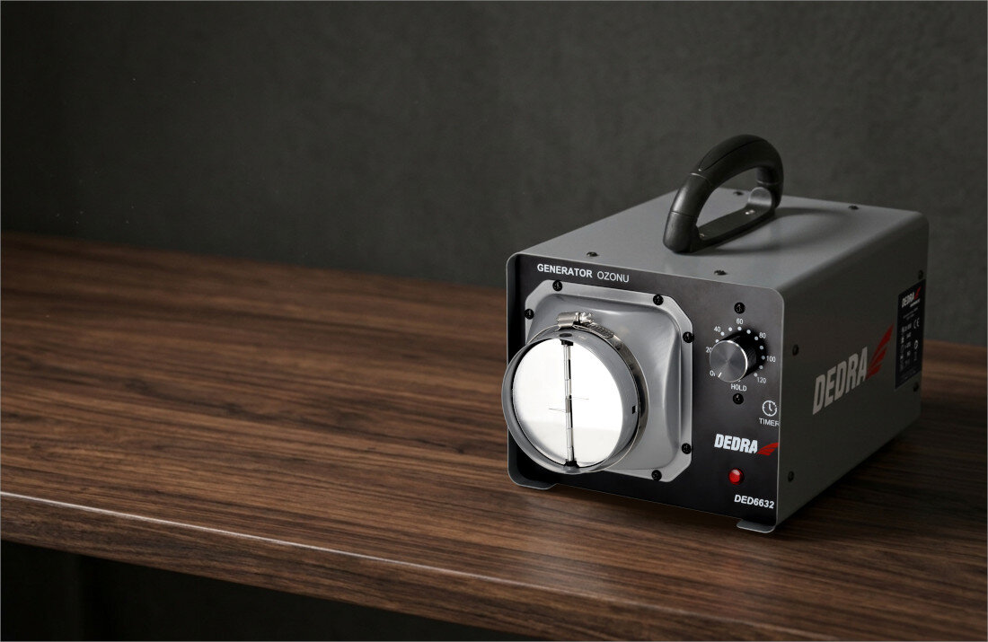 Na drewnianym blacie stoi generator ozonu DEDRA DED6632 z widocznym pokrętłem i uchwytem. Na urządzeniu widnieje napis \'GENERATOR OZONU\' oraz logo \'DEDRA\'.