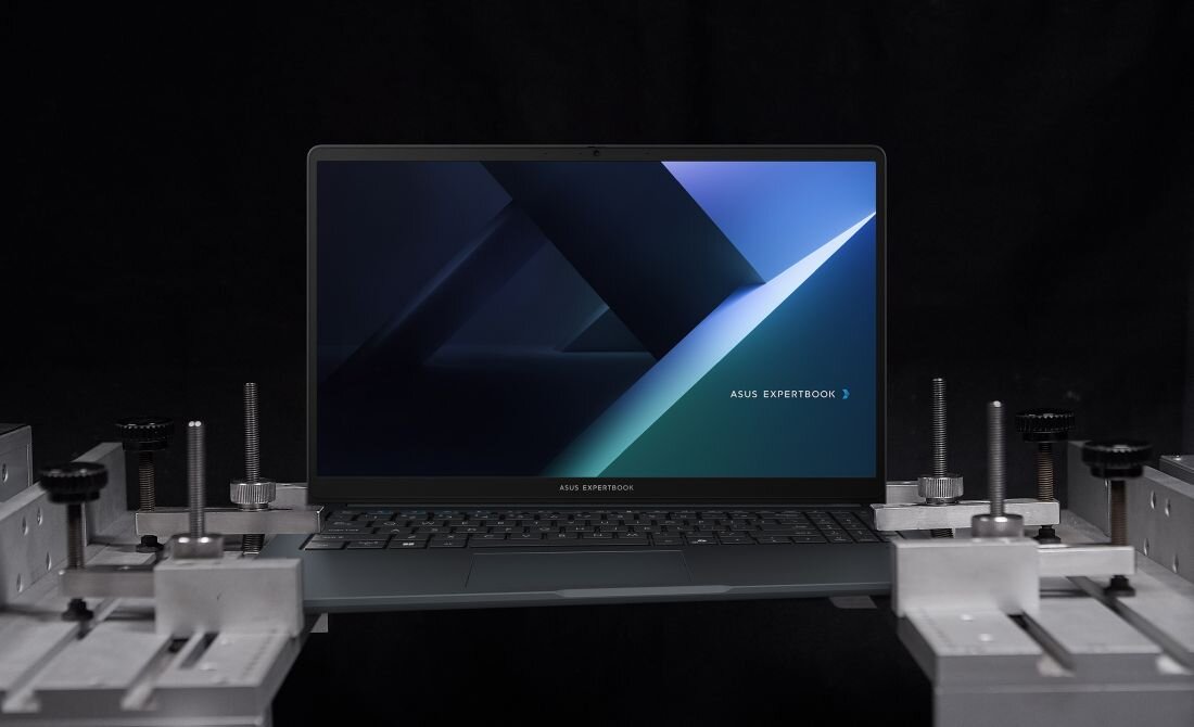 Laptop z wyświetlaczem pokazującym abstrakcyjną grafikę z napisem \'ASUS EXPERTBOOK\'. Urządzenie jest umieszczone na metalowej konstrukcji w ciemnym otoczeniu.