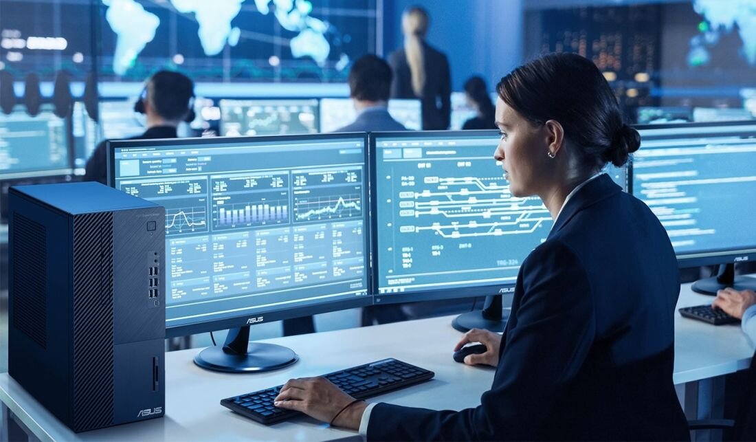Osoba siedzi przy biurku z trzema monitorami, na których wyświetlane są wykresy i dane. Obok monitora znajduje się komputer z widocznym logo ASUS.