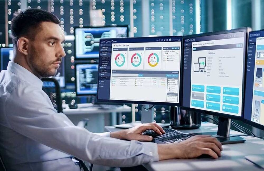 Osoba siedzi przy biurku z trzema monitorami, na których wyświetlane są wykresy i dane systemowe. W tle widać ścianę z migającymi diodami i sprzętem elektronicznym.