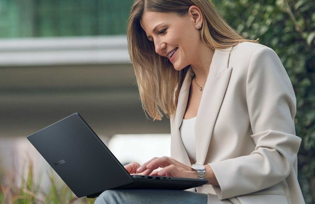 Osoba siedzi na zewnątrz, używając laptopa ASUS. W tle widoczne są rośliny i rozmyte elementy architektury.