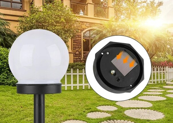 Na obrazie widoczna jest lampa solarna ogrodowa w kształcie kuli, umieszczona na tle ogrodu z trawnikiem i kamienną ścieżką. Obok lampy znajduje się zbliżenie na panel solarny z dwoma pomarańczowymi strzałkami.