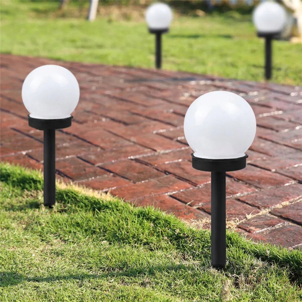 Na obrazie widać kilka kulistych lamp solarnych umieszczonych na trawniku wzdłuż ścieżki z czerwonej cegły. Lampy mają białe klosze i czarne podstawy.