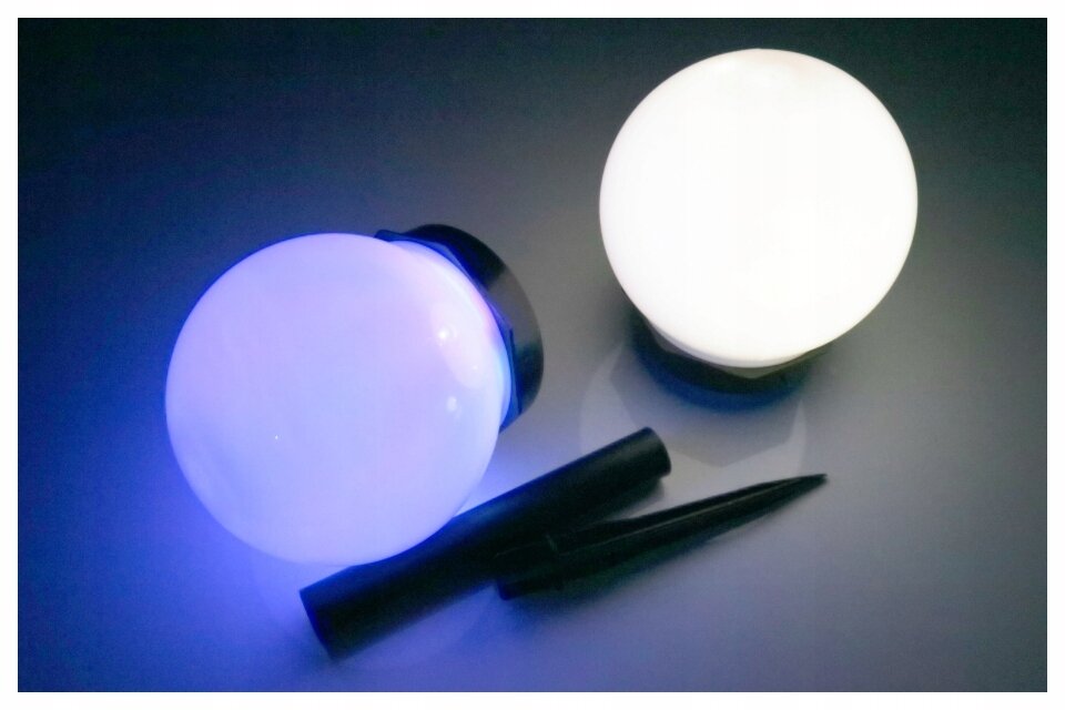 Na obrazie widoczne są dwie kuliste lampy solarne, jedna świeci na niebiesko, a druga na biało. Obok nich leżą dwa czarne elementy przypominające uchwyty lub podstawki.