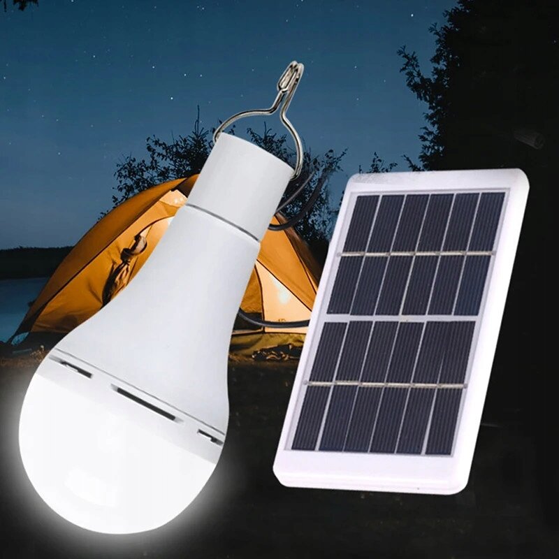 Obraz przedstawia żarówkę LED z uchwytem oraz panel solarny na tle namiotu w nocy. Żarówka jest biała, a panel ma ciemne ogniwa w jasnej ramie.