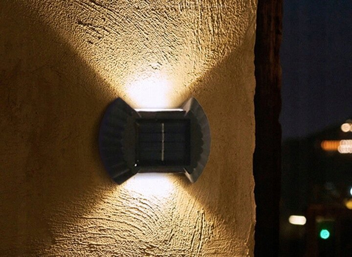 Na ścianie zamontowana jest lampka solarna LED, emitująca światło w górę i w dół. Ściana ma teksturę przypominającą tynk, a w tle widać nocne niebo.
