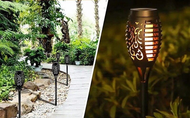 Obraz przedstawia kilka lamp solarnych w kształcie pochodni z efektem płomienia, ustawionych wzdłuż ścieżki w ogrodzie. Lampy mają ażurowe wzory i emitują ciepłe światło przypominające ogień.